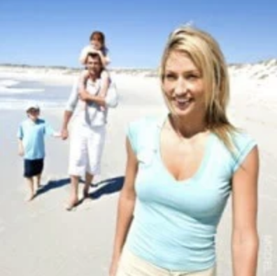 Family at the beach. Father with a child on his shoulders, holding the hand of another young child. Mother walks in front, smiling.