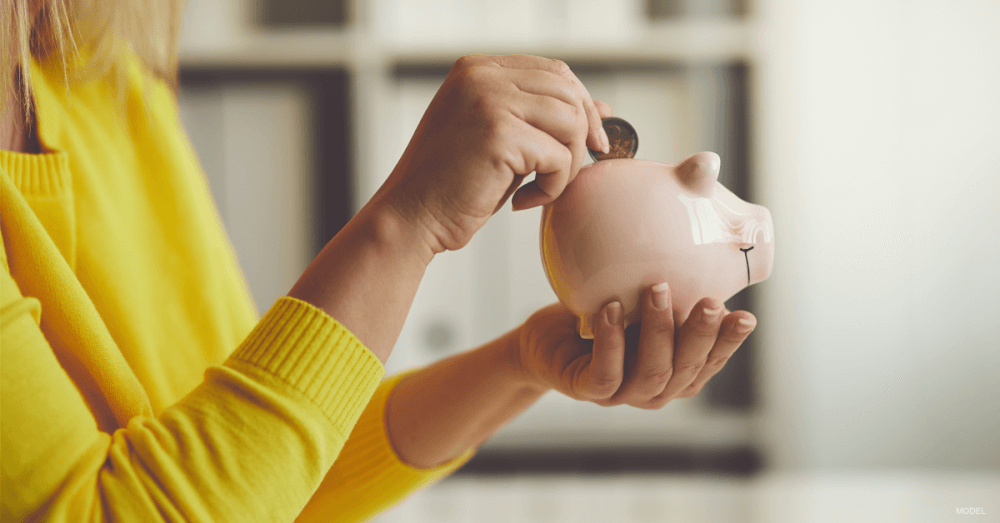 Person wearing a bright yellow sweater, drops a coin into a small, stylized ceramic piggy bank