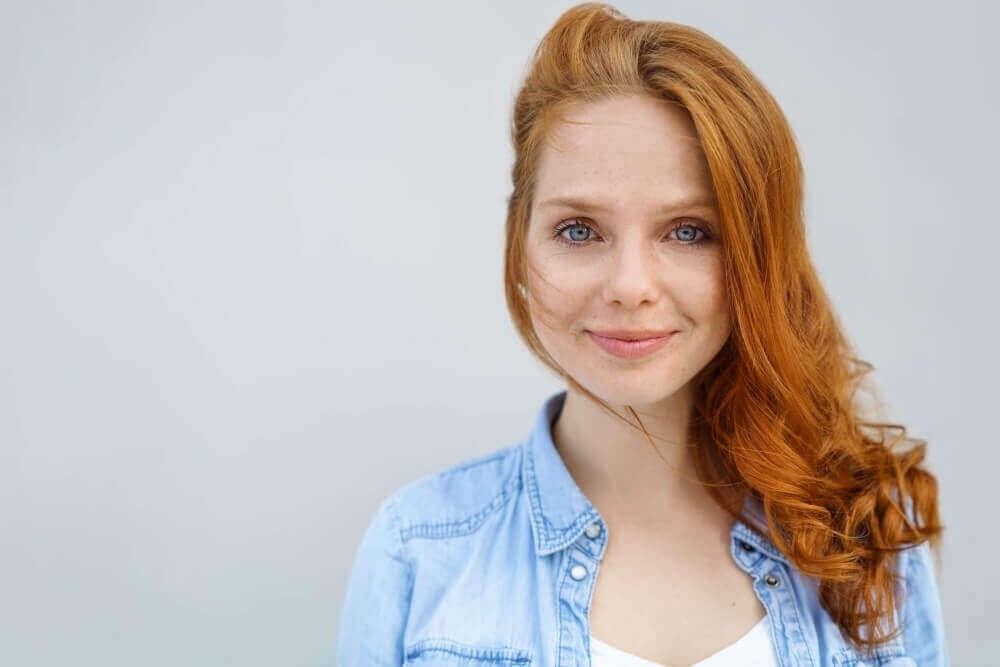 portrait of a woman with red hair, wearing a white tank and open denim shirt, smiling and looking forward
