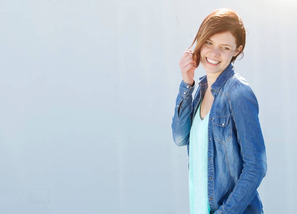 Young woman wearing white shirt and demin jacket, smiles, brushing hair from her face
