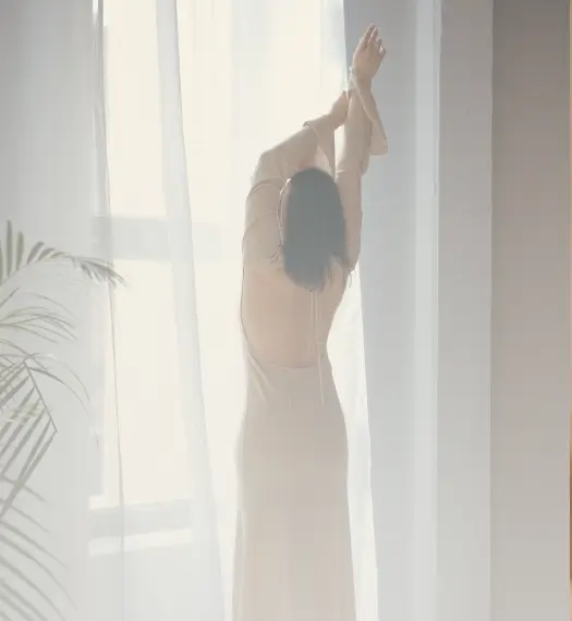 woman stretches her arms up as she looks out a sunlit window